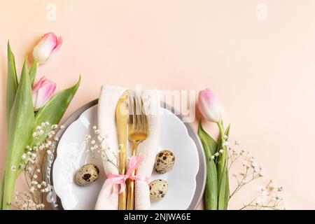 Quail eggs, golden cutlery in napkin on plates, pink tulips and gypsophila on light peach background. Festive composition for Easter table. Spring tab Stock Photo