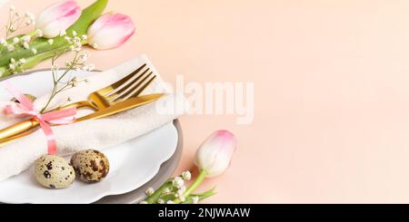 Festive Easter table decoration idea in peach-pink colors. Golden cutlery, quail eggs, napkin, figured wavy plates, pink tulips and gypsophila flowers Stock Photo
