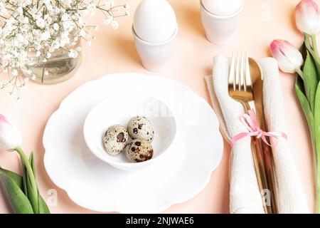 Table setting idea in gentle pink color for festive Easter breakfast or dinner. Eggs on beautiful dishware, golden cutlery, pink tulips and white gyps Stock Photo