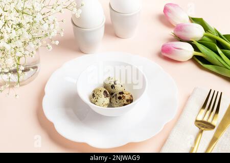 Easter table setting idea. Chicken and quail eggs in egg stands and in beautiful dishware, golden cutlery, pink tulips, white gypsophila. Festive imag Stock Photo