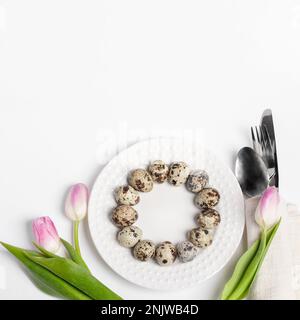 Easter festive layout with quail eggs laid out as circle on plate, pink tulips, silver cutlery on white. Easter table setting idea. Top view. Copy spa Stock Photo