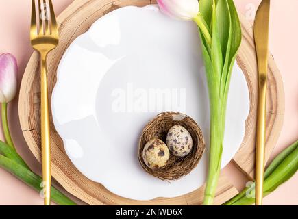 White figured wavy plate, two quail eggs in nest, golden cutlery, pink tulips on round wooden stand. Easter table setting. Festive mock up close up. T Stock Photo