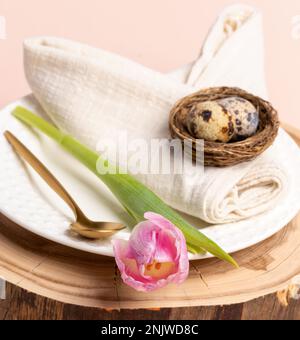 Quail eggs in nest on beautifully rolled up like bunny ears cotton napkin, white plate, golden spoon, pink tulip on round wooden stand. Idea of Pascha Stock Photo