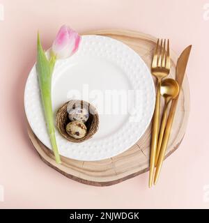 Idea of decoration of Paschal table. Quail eggs in nest on white wavy plate, golden cutlery, tulip on round wooden stand on peach color background. Ea Stock Photo