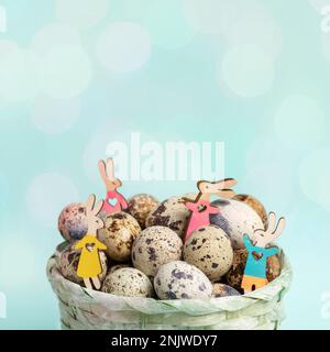 Four small wooden vibrant colored Easter bunnies among quail eggs in light blue woven basket on pastel blue background with bokeh lights. Easter festi Stock Photo