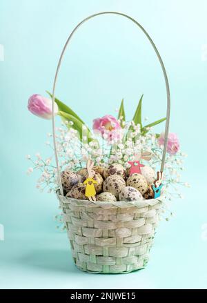 Small wooden vibrant colored Easter bunnies in light blue woven basket with quail eggs, pink tulips and white gypsophila. Easter festive card. Holiday Stock Photo