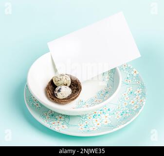 Table setting with two Easter quail eggs in nest, white blank sheet on plates with floral pattern on pastel blue. Romantic Easter congratulation idea. Stock Photo