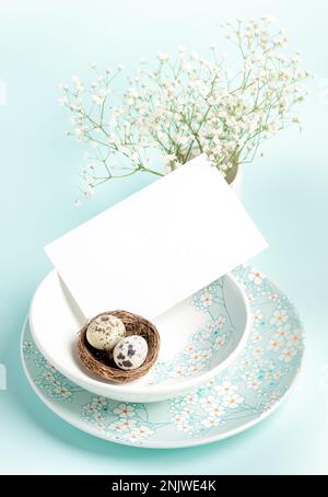 Easter card. Two quail eggs in nest, white empty note, plates with floral pattern on light blue. Romantic Easter. Holiday table setting. Congratulatio Stock Photo