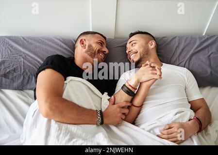 Gay couple looking at each other and smiling while holding hands in bed. Stock Photo