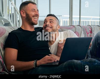 Male couple smiling and having fun together while using a laptop on the sofa. Stock Photo