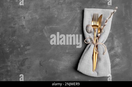 Easter minimal monochrome image. Table setting. Linen napkin with golden cutlery, quail egg, willow twig on gray. Happy Easter holiday concept. Top vi Stock Photo