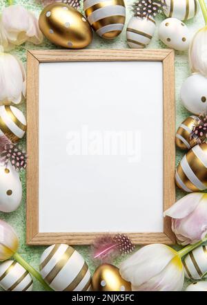 Blank picture frame surrounded by Easter eggs and tulip flowers on green background. Top view, flat lay composition Stock Photo