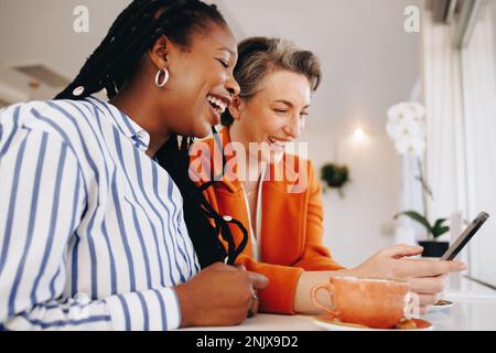 Two female colleagues smiling cheerfully while using a smartphone together in an office cafe. Happy business women enjoying their coffee break togethe Stock Photo