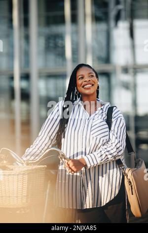 Happy young businesswoman looking away thoughtfully while walking alongside her bicycle in the city. Cheerful young business woman travelling to her o Stock Photo
