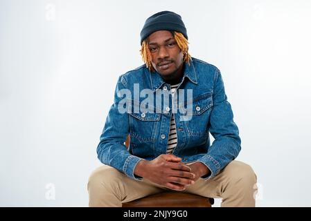 Smiling young African American man with dreadlocks in denim jacket and hat sitting on chair with folded arms and looking at camera on white background Stock Photo