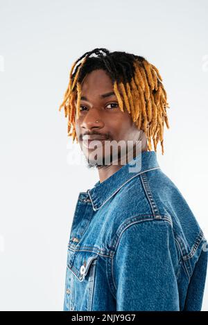 Side view of smiling young African American man with dreadlocks in denim jacket and hat looking at camera on white background Stock Photo