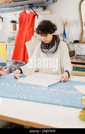 Concentrated female designer in casual clothes and scarf preparing pattern on fabric while working at table in atelier Stock Photo