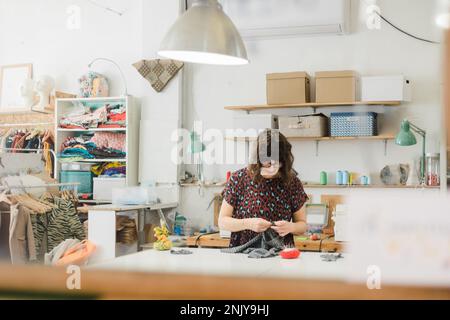 Concentrated female dressmaker in casual clothes and eyeglasses sewing pattern on fabric while working at desk with tools in workshop Stock Photo