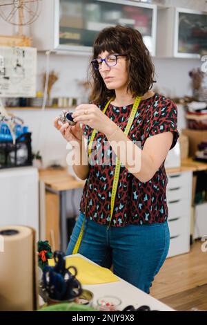 Focused female in casual clothes and eyeglasses standing at table while taking pin needle for creating handmade dress in creative workshop Stock Photo