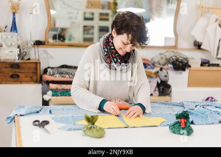 Concentrated smiling female designer in casual clothes and scarf preparing pattern on fabric while working at table in atelier Stock Photo