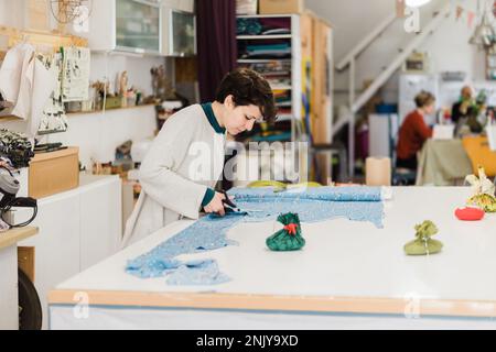 Side view of female tailor in casual sweater cutting piece of colorful fabric with scissors while working on project in creative atelier Stock Photo