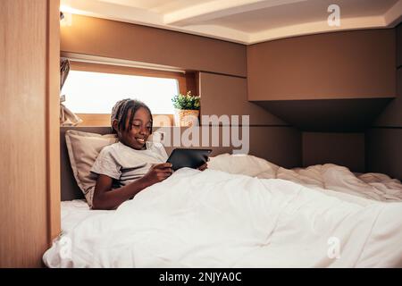 Positive smiling African American preteen boy with dreadlocks lying on comfortable bed and surfing internet via tablet Stock Photo