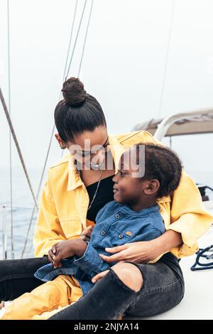 Happy African American mother in casual clothes sitting on yacht while smiling and embracing son during weekend Stock Photo
