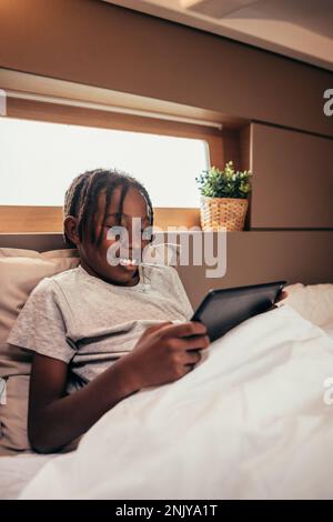 Positive smiling African American preteen boy with dreadlocks lying on comfortable bed and surfing internet via tablet Stock Photo