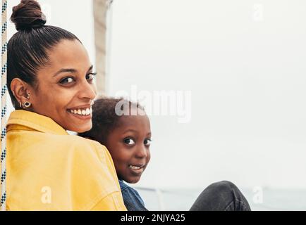 Happy African American mother in casual clothes sitting on yacht while smiling looking at camera and embracing son during weekend Stock Photo