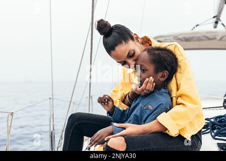 Happy African American mother in casual clothes sitting on yacht while smiling and embracing son during weekend Stock Photo