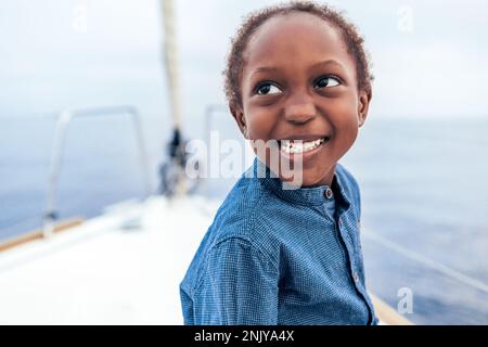 Positive African American boy in casual shirt smiling and looking away while sitting on yacht against sea Stock Photo