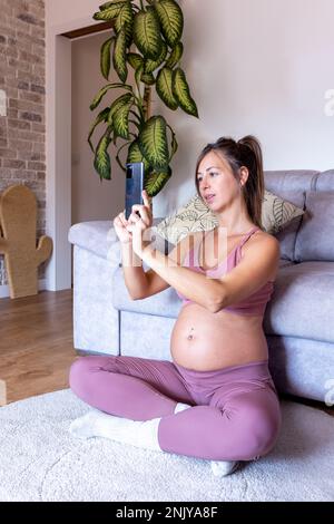 Happy young pregnant female in pink top smiling and taking photo on smartphone while sitting on the floor near sofa at home Stock Photo