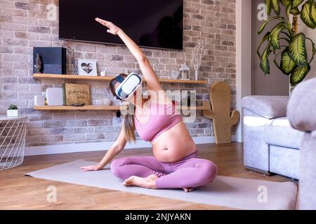 Full body of pregnant female in sportswear sitting on mat in lotus pose and doing side bend with raised arm while practicing yoga in VR headset at hom Stock Photo