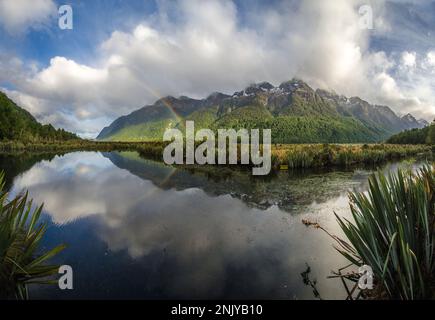 Scenic view of Mirror Lakes reflecting cloudy blue sky and rainbow near green mountains in New Zealand Stock Photo