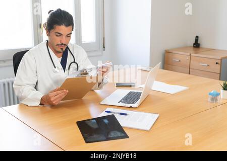 Ethnic young male doctor in white uniform sitting at table with laptop and x ray image while checking information on clipboard in modern hospital Stock Photo