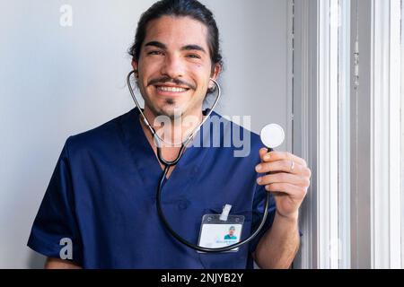 Cheerful ethnic male doctor in blue medical uniform smiling and looking at camera while demonstrating stethoscope against window in hospital Stock Photo