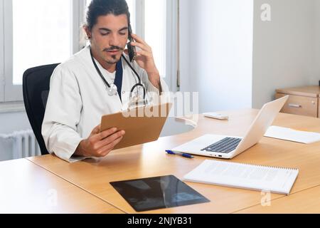 Concentrated male doctor in medical uniform sitting at table with laptop and talking on smartphone while writing notes on clipboard in modern clinic Stock Photo