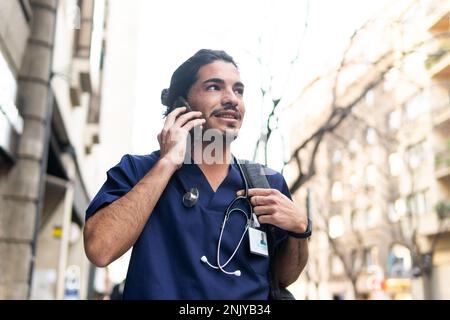 Calm ethnic male doctor in medical uniform talking on cellphone and looking away while walking along city street Stock Photo
