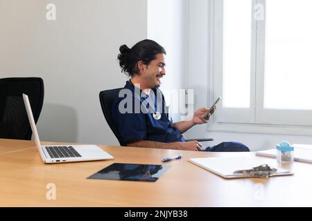 Positive ethnic male doctor in medical uniform with stethoscope smiling and browsing smartphone while sitting at table with laptop during break in cli Stock Photo