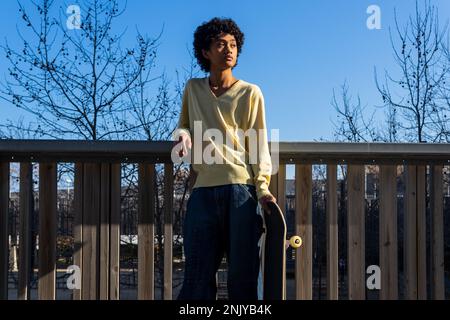 Thoughtful young ethnic male skater in casual outfit with Afro hairstyle looking away while leaning on wooden fence with skateboard against leafless t Stock Photo