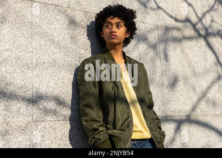 Confident young Asian male with Afro hairstyle in yellow sweatshirt looking away against tiled wall with shadow in sunlight Stock Photo