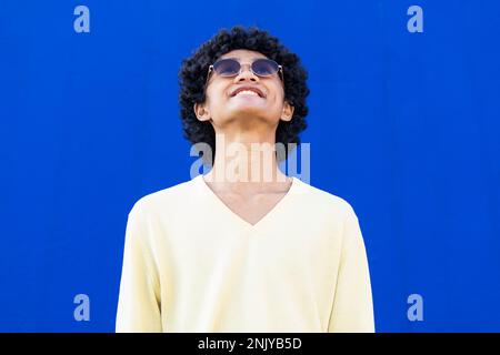 Positive young male in yellow sweatshirt and sunglasses with curly hair looking up and smiling while standing against bright blue wall Stock Photo