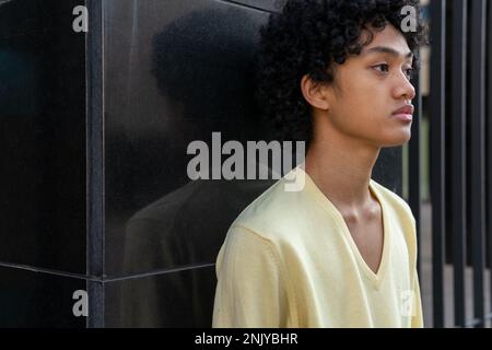 Side view of serious young ethnic male with curly hair wearing yellow sweatshirt looking at camera near black wall on city street Stock Photo