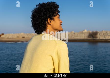 Back view of thoughtful smiling young Asian male skater in yellow sweatshirt with curly hair sitting on embankment enjoying sea Stock Photo