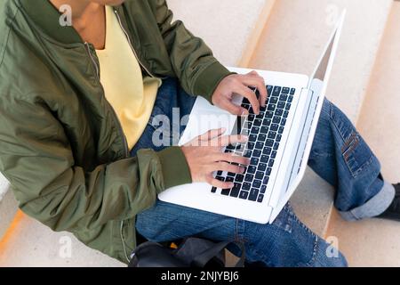 From above cropped unrecognizable young ethnic male student in casual clothes with backpack and Afro hairstyle browsing netbook while doing homework o Stock Photo