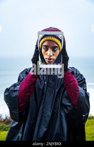 Portrait of cheerful young female traveler in raincoat covering face with digital tablet with self photo on screen against sea under blue sky Stock Photo