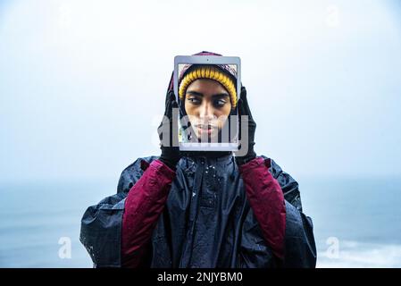 Portrait of cheerful young female traveler in raincoat covering face with digital tablet with self photo on screen against sea under blue sky Stock Photo