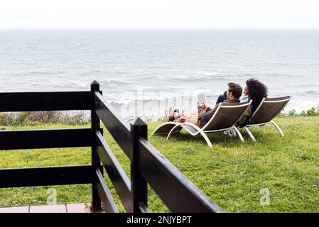 Multiracial girlfriend and boyfriend smiling and using tablet while chilling on deckchairs on grassy shore near stormy sea Stock Photo