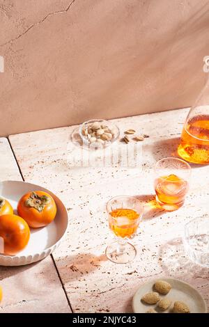 High angle of alcohol cocktail in shot glasses with ice cubes placed on marble tile near persimmon fruits and nuts in sunlight Stock Photo