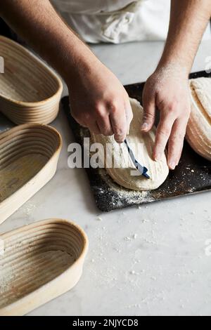 Anonymous baker in apron with blade scoring raw sourdough loaf on table while preparing bread in kitchen Stock Photo